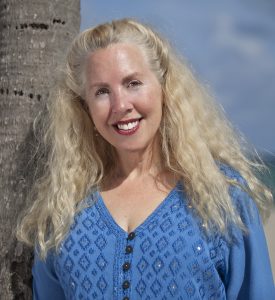 A blonde woman with long curly hair wearing a blue diamond-patterned blouse. She is leaning against a tree.